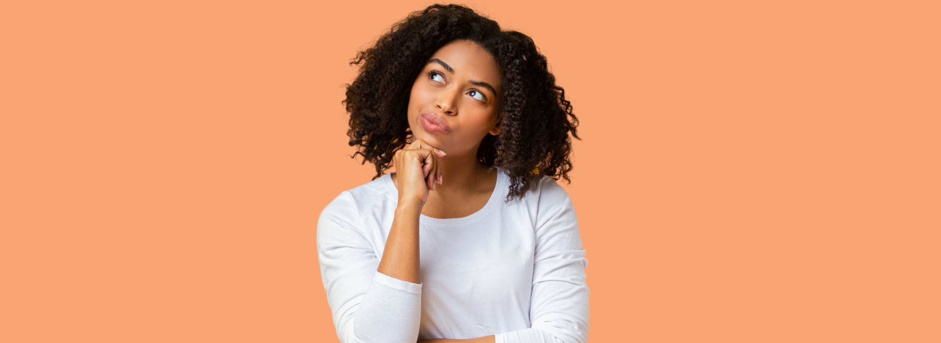 Foto de Capa: Mulher de cabelos cacheados com a mão no queixo olha para cima enquanto está pensativa