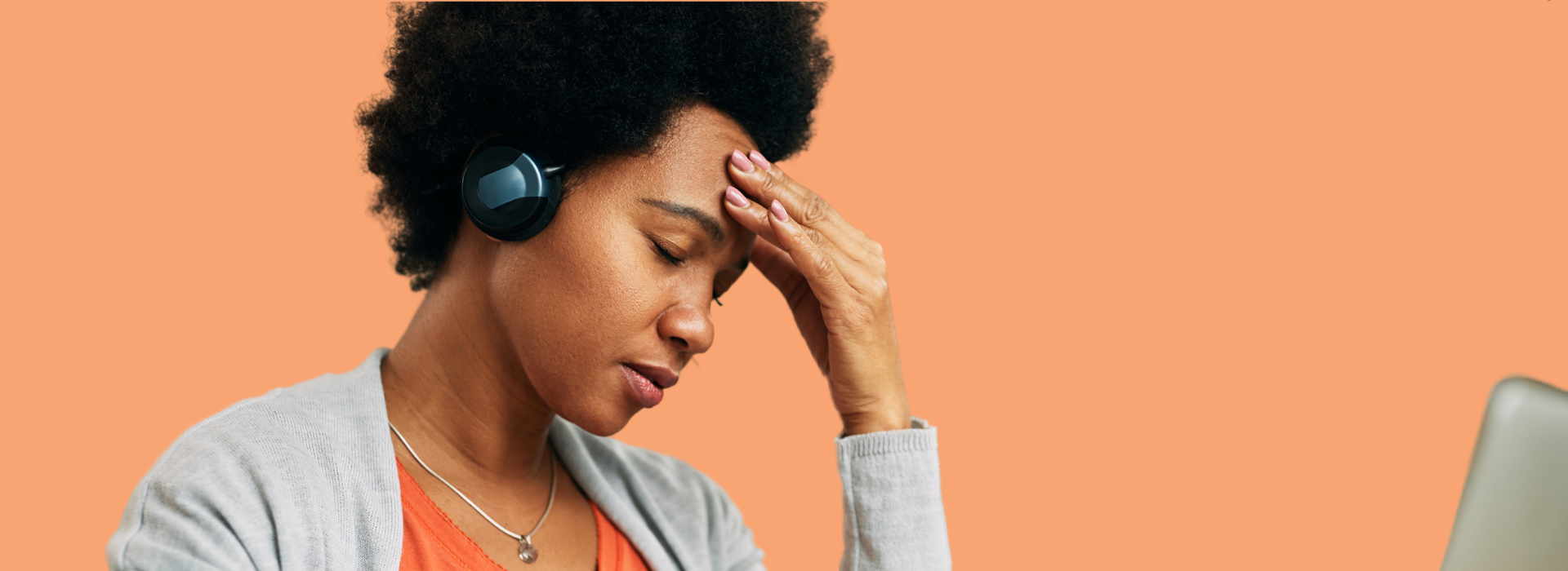 Foto de Capa: Mulher de abafador está de olhos fechados e com uma das mãos na testa enquanto está sentada em frente ao computador