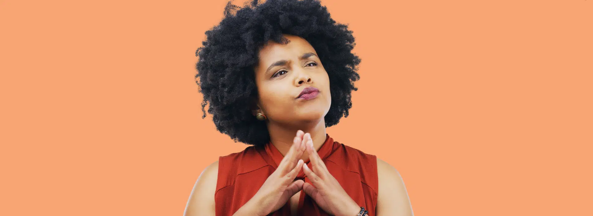 Mulher de cabelos cacheados tem feição instigante enquanto junta as duas mãos