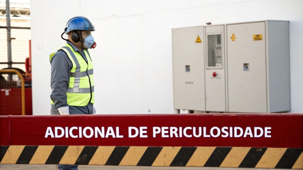 Homem usando equipamentos de proteção individual (capacete, máscara, colete) em ambiente industrial com barreira de segurança.